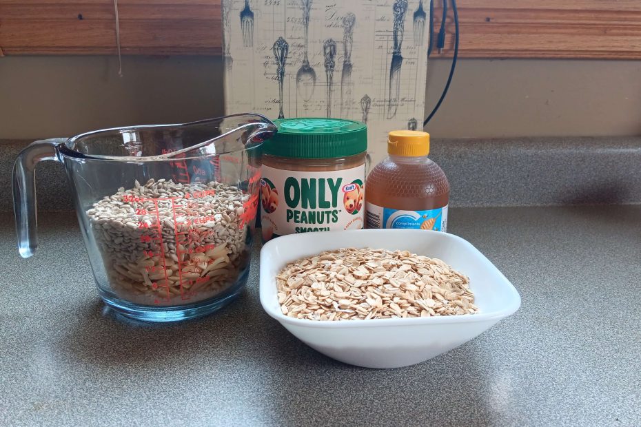 Granola bar ingredients: 2 cups of oats in a bowl, measuring cup holding 3 cups of nuts and seeds, a jar of peanut butter and a bottle of honey.